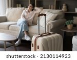 Focus on beige suitcase with blurred carefree young woman sitting on sofa on background. Joyful refreshed millennial Hispanic lady satisfied with vacation travel, relaxing alone in modern hotel room.