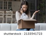 Small photo of Dissatisfied angry woman, cheated client sit on sofa check received box, damaged or broken goods in parcel, talks to customers support, express complaints, looks annoyed. Bad delivery services concept