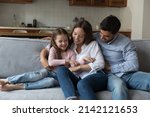 Happy joyful parents, mom and dad cuddling, tickling little daughter kid, sitting, playing on comfortable couch, having fun, enjoying family funny leisure time, parenthood, laughing