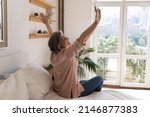 Small photo of Side view older woman sit on bed in modern light warm bedroom raise hands stretch body muscles feels happy, welcomes new day, enjoy carefree morning, untroubled retired life and comfort living concept