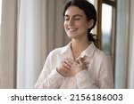 Small photo of Young lovely woman standing alone indoors closed her eyes put folded palms on chest looks grateful, feeling deep appreciation, express sincere feelings. Believe, charity, symbol of gratitude concept