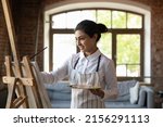 Young beautiful Indian woman painter creates pictures, drawing with paints on canvas in modern workshop. Professional occupation, creative hobby, activity for soul, vocation, develop skills concept