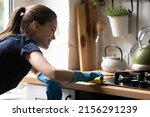 Happy tenant girl using blue protective rubber gloves, rug, cleaning kitchen countertop, cooktop, smiling, enjoying cleanup. Household chores, cleaning service, domestic hygiene concept