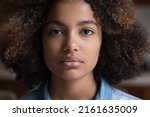 Calm serious beautiful millennial African American woman with thick curly hair looking forward at camera posing indoors. Attractive focused young adult gen z lady face without smile. Close up portrait