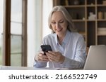 Smiling middle-aged businesswoman using smartphone seated at workplace, distracted from work chatting on internet, read message from client, check calendar. Modern tech for business, agenda concept