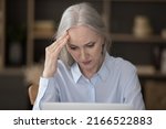 Serious puzzled older businesswoman sit at desk looks at laptop touch head feels stressed due to info overload, faced with difficult task, lack of understanding of tech, new software, bad news concept
