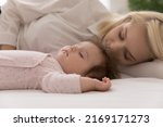 Close up shot peaceful newborn baby and young mom sleeping in bed, lying down with eyes closed on fresh white sheets looking carefree rest together at home. Sweet dreams, protection, maternity concept