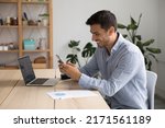 Small photo of Smiling 35s Hispanic businessman, office employee hold cellphone sit at workplace, check e-mail app, usage of mobile business application, comfort easy life use modern wireless tech at work concept