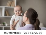 Small photo of Rear back view young loving mom holding in arms her cute newborn, baby looks at camera. Family enjoy playtime together after daytime nap at home. Parent care, mothers love, infancy, motherhood concept