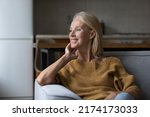 Small photo of Smiling attractive middle-age single woman daydreaming staring out window resting alone seated on sofa in studio apartment. Homeowner portrait, rented flat and tenancy, carefree retired life concept