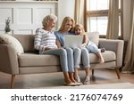 Small photo of Funny movie. Happy family of 3 diverse age female members resting on sofa at home with laptop, hoary aged grandmother, her adult daughter and small grandchild girl watching comedy on computer screen