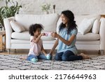 Small photo of African mother and little daughter holding plastic toy teacups enjoy tea ceremony play together sit on carpet in warm living room. Dishware shop advertisement, weekend playtime with children concept