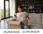 Small photo of Young woman use smartphone unpack received delivered parcel box, takes out baskets, wicker storage jars, pretty things for home interior. Happy client of webstore, e-commerce application usage concept