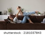 Small photo of Loving mom play with little kid at home, lying on sofa lifts on arms her cute 6s daughter feeling freedom imagining like flying in air. Active playtime leisure, daydreams about family vacation concept
