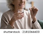 Small photo of Smiling elderly female patient holding pill and glass of water. Senior mature woman taking daily dose of prescribed meds against depression, mental disorders, flu, insomnia, pain. Elderly treatment