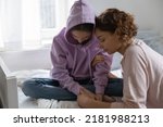 Caring mother hugging, comforting depressed upset teenage daughter, stressed unhappy teenager girl wearing hood sitting on bed, thinking about problems, feeling lonely and misunderstood