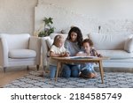 Small photo of Mom teaching cute daughter and son kids to draw in colorful pencils in paper album, sitting at small table on heating floor in living room, enjoying creative process, playtime, family leisure