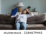 Exhausted young Indian girl feeling hot, tired, sick, resting on couch at home, waving paper handheld fan for cooking, suffering from heat attack, hypoxia, headache, touching head