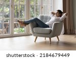 Calm peaceful lazy young Indian homeowner woman resting in armchair with closed eyes at big terrace window, breathing fresh cool air, enjoying peace, leisure, comfort, meditating at home
