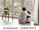 Back view of black haired young mom and little daughter girl sitting on floor at home, hugging with love, care, looking at panoramic window, cozy balcony. Motherhood, real estate concept