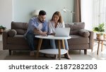 Small photo of Serious millennial couple sit on sofa sorting out papers, reviewing bills for utility expenses, analysing taxes, do household payment use e-bank application on laptop. Manage finances concept