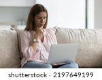 Focused young Caucasian freelance business woman using laptop computer at home, sitting on couch, looking at display, touching chin, thinking, working, watching, reading content