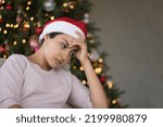 Depressed frustrated Indian girl in Christmas hat going through problems, emotional crisis, negative mood, feeling apathy, loneliness, sitting on sofa at Xmas tree lights, thinking over bad news