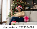 Laughing happy young African woman in festive mood holding glass with sparkling wine in hands resting on comfortable sofa in decorated with Christmas tree living room, having fun at home.