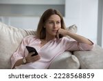 Pensive attractive woman rests seated on sofa at home with mobile phone in hand, feels concerned, waits for call, remember information, recall telephone number, distracted from gadget staring aside