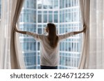 Rear view young woman standing indoors, looking out panoramic window of luxury modern apartment or hotel room opens curtains in morning, enjoying city skyscrapers view, feels happy, welcoming new day