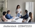 Serious pretty millennial project leader woman talking to employees on meeting, standing at table, explaining tasks, presenting strategy, plan. Female boss mentoring team