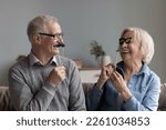 Small photo of Happy joyful older couple of grandparents playing with photo shooting props, applying fake moustache and glasses on sticks to faces, having fun, smiling, laughing