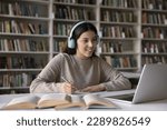 Pretty student teen 17s girl wear headphones e-learns subject, prepare for exams seated at desk studying through videoconference with tutor using laptop in library. Gen Z engaged in on-line education