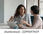 Two business women sit at desk discuss project details, diverse female colleagues met in office, share opinion, working on collaborative task, sales manager makes commercial offer to company client