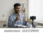 Cheerful excited young Indian blogger recording selfie video on mobile phone, speaking at cellphone fixed on tripod, smiling, laughing, using hand gesture, enjoying self shooting