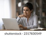 Small photo of Confident young indian woman worker sit at office table by laptop search data browse corporate network for business news. Smiling female freelancer read document online correspond with client in chat