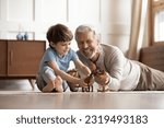Happy elderly grandfather lying on floor at home have fun play toys with small 6s grandson together. Smiling mature grandparent feel playful engaged in funny activity with little boy child on weekend.