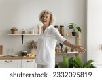 Small photo of Cheerful pretty mature woman having fun at home kitchen, dancing with open arms, laughing, enjoying music, active leisure time, feeling happy, carefree, inspired