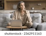 Small photo of Serious thoughtful mature freelance business woman working at laptop at home, using domestic Internet connection, looking away, thinking, making decision, sitting on sofa at computer, touching chin