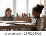 Small photo of Adorable little African girl holding paintbrush drawing pictures on canvas, sit at table with groupmate, attend art classes or daycare, engaged in favourite creative hobby. Talent development of kids