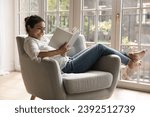 Small photo of Young 25s smiling Indian woman reading novel book relaxing alone on comfort armchair in cozy room, spend leisure enjoy favourite literature bought in bookshop. Booklover pastime at home, hobby concept