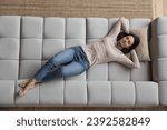 Peaceful young woman in casual clothes relaxing alone on cozy sofa with hands behind head and eyes closed, above view. Fatigue relief, comfortable furniture store ad, carefree leisure at home concept