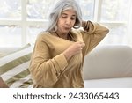 Small photo of Overheated old senior Latin woman blowing under oversized shirt, feeling bad, uncomfortable, hot, suffering from hypoxia, humidity, sweating, heat stroke, sitting on sofa