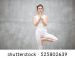 Portrait of beautiful young woman wearing white sportswear working out against grey wall, doing yoga or pilates exercise. Standing in Vrksasana, Tree pose