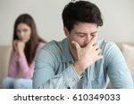 Young guy is very upset and crying, feeling guilty, depressed man in pain, girlfriend sitting in the background indoors, family relationships problems concept, businessman having troubles at work 