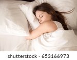 Top view of attractive young woman sleeping well in bed hugging soft white pillow. Teenage girl resting, good night sleep concept. Lady enjoys fresh soft bedding linen and mattress in bedroom 