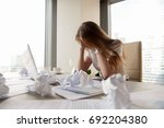 Frustrated businesswoman holding head in hands sitting at office desk covered with crumpled paper, feeling tired after unproductive work, giving up, creative crisis, no motivation, lack of ideas