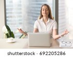 Mindful businesswoman practices breathing exercises at workplace, peaceful woman enjoys yoga with eyes closed at desk, no stress, keep calm, hands in chin mudra gesture, office meditation, portrait