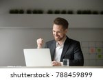 Happy young businessman in suit looking at laptop excited by good news online, lucky successful winner man sitting at office desk raising hand in yes gesture celebrating business success win result