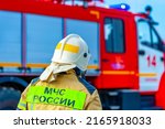 Small photo of Rear view of a Russian firefighter, rescuer in a protective suit and white helmet fighting a fire. Fire extinguishing operation. Behind a sign with the words "EMERCOM of Russia"